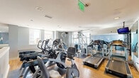 Located in the heart of downtown Dallas at 1509 Main Street, this fitness facility offers a thoughtfully designed single-floor layout divided into two distinct functional areas. The main exercise room is outfitted with a comprehensive selection of cardio machines, including Cybex ARC trainers, ellipticals, stationary bikes, and treadmills, all positioned atop warm hardwood flooring. A full wall of mirrors amplifies the sense of space and supports proper form during workouts, while a mounted flat-screen television adds convenience for users. Natural light streams in through multiple windows, offering views of the surrounding Dallas skyline. The space is finished with soft blue-gray walls accented by a striking geometric mural, creating an energizing yet focused atmosphere. An adjacent studio area provides an open, flexible space ideal for stretching, yoga, or functional training. This room features the same rich hardwood floors, floor-to-ceiling mirrors with sliding glass partitions, and large windows framing downtown Dallas cityscapes. Accessories including medicine balls, kettlebells, and resistance equipment are neatly organized throughout. Recessed lighting and clean white ceilings complete the polished, modern aesthetic of this well-appointed urban fitness space.