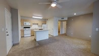 This single-floor unit is designed around an open-concept layout that seamlessly connects the kitchen and living area, creating a functional and cohesive living space. The kitchen is outfitted with light wood cabinetry, a white appliance suite including a range, microwave, refrigerator, and dishwasher, and a central island that provides additional prep and counter space. The flooring transitions from tile in the kitchen to plush carpet in the living area, defining each zone while maintaining an open feel. A ceiling fan with an integrated light fixture is centrally positioned overhead, contributing to both air circulation and ambient lighting alongside recessed lighting. Neutral-toned walls and light-colored finishes give the space a clean, understated aesthetic. A hallway visible from the main living area leads to additional rooms, including a bathroom, indicating a practical and well-organized floor plan. HVAC vents are integrated into the ceiling for climate control throughout. The overall ambiance is straightforward and functional, offering a move-in-ready environment with a cohesive neutral palette.