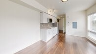 Located at 5001 Brooklyn Ave NE in Seattle's University District, this efficiently designed studio unit makes smart use of its compact footprint across a single floor. The open-concept kitchen and living area serves as the heart of the space, featuring sleek white cabinetry extending to the ceiling, a tiled backsplash, stainless steel appliances including a built-in microwave and refrigerator, and a clean white countertop with an undermount sink. Warm hardwood-style flooring runs throughout the main living area, complementing the bright, neutral white walls and creating a cohesive, modern aesthetic. Track lighting illuminates the kitchen and living space, while large windows allow natural light to filter in, offering views of the surrounding greenery. The entrance provides a practical transition point into the unit, with the bathroom conveniently accessible nearby. The bathroom features white tile flooring, a modern vanity with a mirror, a toilet, and a walk-in shower, with a frosted window providing privacy while still admitting natural light. A dedicated closet offers additional storage. The overall ambiance is clean, minimalist, and contemporary, well-suited for efficient urban living in the heart of Seattle.