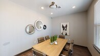 This single-floor conference room is a well-appointed meeting space designed for focused collaboration and small group gatherings. At its center sits a rectangular natural wood table surrounded by cushioned leather chairs in a warm taupe tone, offering comfortable seating for up to eight participants. The room features dark hardwood-style flooring that contrasts cleanly with soft gray walls, creating a professional yet inviting atmosphere.

One wall serves as a distinctive focal point, displaying a cluster of three decorative gear-shaped mirrors in varying sizes and finishes — a silver, a smaller silver, and a dark metal tone — lending the space an industrial-modern character. On the adjacent wall, a large framed portrait of a dog adds a playful and approachable artistic touch.

Natural light filters into the room through a wide window fitted with horizontal blinds, offering a view of the surrounding exterior. A glass-paneled door provides additional access to adjacent areas. Recessed ceiling lighting supplements the natural light, while a centrally mounted ceiling fixture anchors the room overhead. A live snake plant centerpiece on the table introduces a natural, refreshing element. The overall design balances functionality and personality, making it a practical and visually engaging space for meetings and discussions.