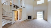 This two-floor space makes an immediate impression with its dramatic, soaring ceilings that create an expansive sense of openness in the main living area. A sleek white metal spiral staircase serves as both a functional and architectural centerpiece, connecting the two levels while adding a striking visual element to the room. The upper level features a balcony-style overlook with matching white metal railings, adding depth and dimension to the vertical space.

The main living area is finished with wall-to-wall carpet in a neutral tone, offering a warm and comfortable underfoot feel throughout. A tile-surround fireplace anchors one corner of the room, providing a cozy focal point ideal for relaxing. Large sliding glass doors on the opposite wall flood the space with natural light and offer direct access to an outdoor balcony or patio, seamlessly blending interior and exterior living.

The lower level also includes additional rooms accessible through interior doorways, suggesting dedicated spaces for sleeping, storage, or utility. The overall color palette of soft whites and warm neutrals lends the space a clean, airy ambiance. High clerestory windows near the roofline further enhance natural light penetration, making the interior feel bright and welcoming throughout the day.