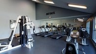 This fitness center offers a clean, contemporary workout environment designed to support a full range of exercise routines. The space features a dark, rubberized floor that provides both durability and safety during high-intensity training sessions. The walls are finished in a deep charcoal gray, lending the room a focused and energetic atmosphere.

A row of treadmills serves as the centerpiece of the cardio section, positioned in front of a large floor-to-ceiling mirror that visually expands the space and allows users to monitor their form during workouts. Overhead fluorescent lighting ensures the area is well-lit throughout. A multi-use strength training machine is positioned to one side, offering a variety of resistance exercises, while additional cardio equipment, including a stationary bike, rounds out the selection.

The motivational phrase "JUST DO IT." is displayed prominently on the upper portion of the wall above the mirror, reinforcing an energetic and driven workout environment. The layout is thoughtfully arranged to maximize floor space and allow for comfortable movement between equipment. Overall, this fitness center combines functional equipment, smart design, and an inspiring aesthetic to create an effective and inviting space for exercise.
