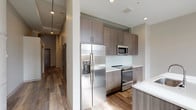 This single-floor unit showcases a thoughtfully designed layout that blends modern finishes with functional living spaces. The kitchen serves as a standout feature, outfitted with textured wood-grain cabinetry extending floor to ceiling, crisp white quartz countertops, a white subway tile backsplash, and a full suite of stainless steel appliances including a French door refrigerator with ice maker, electric range, and built-in microwave. A peninsula with matching countertop provides additional workspace and casual seating.

The open living area adjoins the kitchen, featuring light wood-look plank flooring and tall windows that invite natural light. The exposed concrete ceiling detail adds an industrial character to the space. The bedroom offers a quieter atmosphere with plush carpeting, a ceiling fan, and a high clerestory window for natural light while maintaining privacy.

A dedicated laundry area accommodates a stacked front-load washer and dryer with wire shelving for organization. The bathroom is finished with large-format tile flooring, a walk-in glass-enclosed shower with a rainfall-style fixture, a wood-grain vanity with white countertop, and clean neutral walls throughout, creating a calm and cohesive aesthetic.