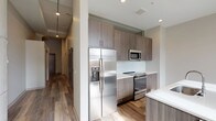 This single-floor unit showcases a thoughtfully designed layout that blends modern finishes with functional living spaces. The kitchen serves as a standout feature, outfitted with textured wood-grain cabinetry extending floor to ceiling, crisp white quartz countertops, a white subway tile backsplash, and a full suite of stainless steel appliances including a French door refrigerator with ice maker, electric range, and built-in microwave. A peninsula with matching countertop provides additional workspace and casual seating.

The open living area adjoins the kitchen, featuring light wood-look plank flooring and tall windows that invite natural light. The exposed concrete ceiling detail adds an industrial character to the space. The bedroom offers a quieter atmosphere with plush carpeting, a ceiling fan, and a high clerestory window for natural light while maintaining privacy.

A dedicated laundry area accommodates a stacked front-load washer and dryer with wire shelving for organization. The bathroom is finished with large-format tile flooring, a walk-in glass-enclosed shower with a rainfall-style fixture, a wood-grain vanity with white countertop, and clean neutral walls throughout, creating a calm and cohesive aesthetic.