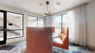 Located at 1724 Partnership Way in Houston, TX, this compact yet thoughtfully designed lounge and meeting room delivers a striking contemporary aesthetic. The space is anchored by a central round table surrounded by high-back orange upholstered booth seating, creating an intimate and functional gathering area. An eye-catching multi-arm sputnik-style chandelier hangs overhead, providing warm, directional lighting that complements the bright, white ceiling. One wall features a bold teal geometric relief panel, serving as a sophisticated focal point that adds texture and visual depth to the room. The patterned carpet, featuring an abstract mix of grays, blues, and whites, adds a dynamic energy underfoot. Large black-framed windows wrap around the space, flooding it with natural light and offering open views of the surrounding outdoor community. Sheer white curtains frame the windows softly. A wall-mounted television above a sleek console table with decorative accents provides additional functionality. A glass-panel sliding door connects to an adjacent stairwell, enhancing the sense of openness. Overall, the space balances modern design details with practical amenities, making it well-suited for collaborative meetings or casual social gatherings.
