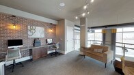 This single-floor space combines industrial and contemporary design elements to create a functional and visually engaging environment. A standout feature is the exposed brick accent wall, which anchors a built-in workstation complete with dark wood cabinetry, a desktop computer, and an ergonomic chair — making it well-suited for focused work or productivity tasks. A warm Edison-style pendant light adds character above the desk area, complementing the textured brick backdrop and a piece of abstract wall art.

The lounge area to the right features a striped, earth-toned upholstered chair, offering a comfortable seating option. Track lighting runs along the ceiling, providing adjustable, directional illumination throughout the space. Large windows along the far wall allow natural light to filter in, brightening the room and offering views to the exterior.

Neutral gray carpeting grounds the space, while the warm taupe wall tones and exposed ductwork contribute to an urban, loft-inspired aesthetic. The open layout and thoughtful mix of work and lounge zones make this a versatile, welcoming space suited for both collaboration and individual use.