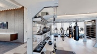 This expansive, contemporary fitness center is designed to deliver a premium workout experience across a thoughtfully organized single-level layout. Floor-to-ceiling glass walls flood the interior with natural light and frame striking views of an outdoor pool and surrounding cityscape, creating an open and energizing atmosphere throughout.

The facility is comprehensively equipped to support a wide range of training styles. A dedicated strength training zone features Matrix cable machines and resistance stations, while a cardio section houses treadmills, ellipticals, and Assault Fitness curved treadmills. For functional and high-intensity training, the space includes a TRX suspension system, kettlebells, medicine balls, and battle ropes neatly organized on a large multi-tier storage rack. Torque Fitness heavy bags are suspended for boxing and combat training sessions.

A distinctive lounge area provides a comfortable retreat adjacent to the workout floor, furnished with sleek white sofas and hexagonal ottomans arranged around a textured wood feature wall with a mounted flat-screen TV. Architectural detailing, including a coffered ceiling with warm recessed lighting and a slatted wood ceiling element, adds a refined, boutique-hotel aesthetic. Light stone tile flooring runs throughout, unifying the space and maintaining its clean, modern character.