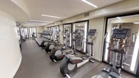 This fitness center is a thoughtfully designed, single-floor facility built to support a well-rounded workout experience. The space is outfitted with a generous lineup of cardio equipment, including treadmills, elliptical trainers, and stationary bikes, all arranged in an organized row for easy access and comfortable use. A continuous bank of large mirrors lines one wall, creating a sense of expanded space while allowing users to monitor their form during exercise. The mirrors are framed with bright, integrated lighting panels that provide even, flattering illumination throughout the room. Overhead, recessed linear lighting runs the length of the ceiling, ensuring the entire floor is well-lit without harsh shadows. The flooring is a durable, fitness-appropriate surface suited for both machine use and free movement. Neutral wall tones and a clean, uncluttered aesthetic give the space a professional and focused atmosphere. Ventilation is addressed with ceiling-mounted HVAC vents, contributing to a comfortable environment during intense workouts. The overall design prioritizes functionality and clarity, making it a practical and welcoming facility for daily fitness routines.
