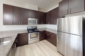 This 890-square-foot, single-floor apartment offers a practical and modern layout with 2 bedrooms, 2 bathrooms, a combined kitchen and living room, in-unit laundry, and a dedicated closet. The kitchen is equipped with stainless steel appliances — including a refrigerator, range, and microwave — complemented by rich dark-espresso cabinetry, granite-style countertops, and light wood-style flooring that extends throughout the main living areas. The open connection between the kitchen and living room creates a cohesive, functional common space filled with natural light. Both bedrooms feature neutral carpet flooring and closet storage, with the primary bedroom measuring 140 square feet. One bedroom benefits from a large picture window offering views of the surrounding neighborhood. Both bathrooms are finished with the same consistent design palette, each featuring a full tub-shower combination, a dark-cabinet vanity with granite countertops, and light wood-style flooring. The in-unit laundry room houses a full-size side-by-side washer and dryer with wire shelving above for additional storage. Throughout the unit, neutral wall tones, clean lines, and consistent finishes create a bright, move-in-ready living environment.