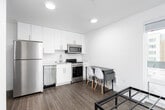 This single-floor studio unit offers a thoughtfully designed open-concept layout that efficiently combines living, sleeping, and dining functions within one cohesive space. The interior features wood-look plank flooring throughout, white walls, and recessed lighting, creating a clean and contemporary aesthetic.

The kitchen is well-appointed with stainless steel appliances, including a full-size refrigerator, electric range, built-in microwave, and dishwasher. White flat-panel cabinetry extends to the ceiling, maximizing storage, while a compact countertop and sink complete the functional workspace. A small dining table with modern chairs is positioned adjacent to the kitchen area.

The sleeping area is defined by large windows that frame views of the surrounding urban streetscape and neighboring buildings, allowing abundant natural light to fill the space. The bathroom maintains the unit's clean design language, featuring a white vanity with an undermount sink, chrome fixtures, a towel bar, and consistent wood-look flooring.

Overall, the unit presents a minimalist, move-in-ready environment well-suited for individuals seeking a practical and modern urban living space.