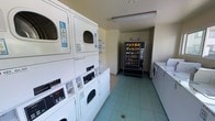 This shared laundry facility offers a practical and well-organized space designed to meet the everyday needs of its users. The room features a row of stacked washer and dryer units lining one wall, maximizing capacity within the available floor space. Additional standalone washing machines are positioned along the opposite side, providing ample options during peak usage times. Each machine is clearly labeled and equipped with digital controls and payment interfaces, allowing for straightforward, self-service operation. A snack vending machine is conveniently located at the far end of the room, offering refreshments while waiting for laundry cycles to complete. The space is finished with light-colored tile flooring and neutral walls, creating a clean and functional environment. Overhead fluorescent lighting ensures the room is well-lit throughout. Informational notices and usage instructions are posted on the walls, helping users navigate the facility with ease. The overall layout is efficient and thoughtfully arranged, making this laundry room a highly practical shared amenity.