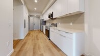 The Haven is a thoughtfully designed single-floor unit that makes efficient use of its compact footprint across a kitchen, living room, dining room, and bathroom. Light-toned hardwood flooring runs continuously throughout the space, lending warmth and visual cohesion to each room.

The galley-style kitchen is a standout feature, outfitted with sleek white flat-front cabinetry extending from floor to ceiling, a neutral tile backsplash, light quartz countertops, and a full suite of stainless steel appliances including a refrigerator, dishwasher, microwave, and range. Recessed lighting illuminates the kitchen corridor, which flows toward a glass exterior door admitting natural light.

The living and dining areas offer clean, white walls and the same warm wood flooring, providing a bright, open canvas. Windows fitted with blinds allow for controlled natural light throughout. The bathroom is spacious relative to the overall unit, featuring a white vanity with a light countertop, wood-look flooring, and a stacked washer/dryer unit conveniently integrated into the space.

Overall, The Haven presents a modern, minimalist interior with a neutral palette, practical layout, and quality finishes that maximize comfort and functionality within a cozy, well-appointed space.