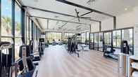 This well-appointed fitness center offers a generously sized, single-floor layout designed to accommodate a comprehensive range of workout activities. The space is characterized by its contemporary architectural design, featuring coffered ceilings with large ceiling fans, exposed dark structural beams, and recessed lighting that complement the bright, airy atmosphere throughout.

Floor-to-ceiling black-framed windows line the perimeter walls, flooding the interior with abundant natural light and offering expansive views of the surrounding landscaped grounds, palm trees, and adjacent water features. The light-toned hardwood-style flooring adds warmth and continuity across the entire space.

The main workout floor is equipped with a diverse selection of strength training machines, including cable systems, a multi-functional cable tower, free weights with a dedicated dumbbell rack, a power rack with bench, and a heavy bag for boxing. Cardio equipment includes treadmills, stair climbers, and elliptical machines, each strategically positioned to take advantage of the outdoor views.

A secondary area serves as a wellness support station, featuring a built-in wood-accented counter with open shelving, a compact refrigerator, and a decorative accent wall with subtle patterned wallcovering. Glass partition walls and sliding glass doors connect the zones, maintaining an open, cohesive feel throughout the facility.