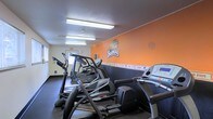 This fitness center is a purpose-built workout facility proudly showcasing Oregon State University Beavers branding throughout. The space is immediately recognizable by its striking orange accent wall, emblazoned with the iconic Beavers logo and the "OS" monogram, creating an energetic and motivating atmosphere for users.

The room is outfitted with a variety of cardio equipment, including elliptical machines, stair climbers, and treadmills, all arranged to allow comfortable circulation and access. The dark flooring provides a practical, durable surface suited for high-traffic athletic use and contrasts boldly with the vibrant orange and neutral-toned walls.

Overhead, fluorescent lighting ensures the space is well-lit throughout, supporting safe and effective workouts at any time of day. The ceiling is finished with a textured surface, and the room maintains a clean, functional layout. A clearly marked exit door is visible at the far end, emphasizing safety and accessibility.

A decorative banner strip runs along the mid-wall, adding additional team spirit and visual interest. The overall ambiance is energetic and school-spirited, making this facility not just a place to exercise, but a space that fosters community pride and athletic motivation for its users.