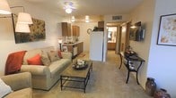 This single-floor residence offers an open-concept layout that connects the living, dining, and kitchen areas into one cohesive space. The living area is comfortably proportioned, featuring plush carpeted flooring and a ceiling fan with integrated lighting that contributes to the warm, inviting ambiance. A cushioned sofa with decorative accent pillows and a wood-and-metal coffee table anchor the seating area, complemented by a small dining table nearby.

The kitchen flows directly from the living space and is equipped with wood-toned cabinetry, a white refrigerator, dishwasher, and ample counter space. A small decorative rug adds a touch of color near the kitchen's base. The cabinetry extends above for generous storage, and the layout allows for easy movement between cooking and entertaining areas.

A hallway visible from the main living space leads to additional rooms, including what appears to be a bathroom with a vanity and mirror. The warm lighting throughout the space, combined with the neutral color palette of beige tones and warm wood accents, creates a comfortable and functional environment suited for everyday living.