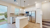 Located at 1126 Green Cypress Loop in Davenport, Florida, this two-story home showcases a clean, contemporary design with a bright and airy aesthetic carried consistently throughout both levels. Light wood-look flooring flows seamlessly across the main living areas, complementing the crisp white walls and recessed lighting that illuminate each space.

The open-concept main level features a generous living area with a tray ceiling detail and a sliding glass door that invites natural light and outdoor views. The kitchen is well-appointed with white shaker cabinetry, a neutral tile backsplash, a center island with a white countertop, and a full suite of stainless steel appliances including a refrigerator, range, microwave, and dishwasher. Pendant lighting above the island adds a refined touch, while a window above the sink brings in additional natural light.

The upper level continues the home's cohesive design, featuring a spacious primary bedroom with a tray ceiling, dual windows, and generous proportions. Additional bedrooms are bright and well-sized with light flooring and window blinds. Bathrooms are finished with white tile surrounds and brushed gold fixtures, adding a modern, polished accent to the overall design.
