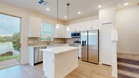Located at 1126 Green Cypress Loop in Davenport, Florida, this two-story home showcases a clean, contemporary design with a bright and airy aesthetic carried consistently throughout both levels. Light wood-look flooring flows seamlessly across the main living areas, complementing the crisp white walls and recessed lighting that illuminate each space.

The open-concept main level features a generous living area with a tray ceiling detail and a sliding glass door that invites natural light and outdoor views. The kitchen is well-appointed with white shaker cabinetry, a neutral tile backsplash, a center island with a white countertop, and a full suite of stainless steel appliances including a refrigerator, range, microwave, and dishwasher. Pendant lighting above the island adds a refined touch, while a window above the sink brings in additional natural light.

The upper level continues the home's cohesive design, featuring a spacious primary bedroom with a tray ceiling, dual windows, and generous proportions. Additional bedrooms are bright and well-sized with light flooring and window blinds. Bathrooms are finished with white tile surrounds and brushed gold fixtures, adding a modern, polished accent to the overall design.