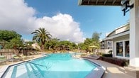 This community amenity space centers around a generously sized, geometric resort-style swimming pool with crystal-clear blue water, designed to accommodate both leisurely swimming and relaxation. The pool's unique angular shape adds a modern design element to the outdoor environment, complemented by a separate spa area finished with decorative tile. Surrounding the pool, a spacious sun deck is outfitted with multiple green lounge chairs, offering ample space for sunbathing and outdoor relaxation. A covered pergola area with umbrella-shaded seating provides a comfortable retreat from the sun. The entire pool area is enclosed by a wooden fence, ensuring privacy and safety. Towering mature palm trees and a canopy of established shade trees frame the space, creating a tropical and serene atmosphere. Adjacent to the pool, a single-story clubhouse building features large windows with views of the pool area, white columns, and a welcoming entrance with integrated landscaping beds. The building's exterior showcases neutral siding with classic architectural detailing. The overall ambiance is bright, open, and resort-inspired, making this outdoor amenity space an inviting focal point of the community.