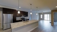 This single-floor residence showcases a thoughtfully designed open-concept layout that seamlessly connects the kitchen and living areas. The kitchen is a standout feature, anchored by an expansive granite-topped island with an undermount sink and elegant pendant lighting that illuminates the workspace below. Rich, dark espresso cabinetry lines the kitchen walls, providing generous storage, while stainless steel appliances — including a range with overhead ventilation — add a sleek, contemporary touch. The kitchen island serves as a natural gathering point and transitions effortlessly into the open living area beyond. Durable wood-look flooring extends throughout the space, adding warmth and continuity to the overall design. The living area is bathed in natural light from a window and is enhanced by a ceiling fan, contributing to a comfortable and airy ambiance. Neutral gray walls throughout create a clean, versatile backdrop that complements the modern finishes. The open sight lines between the kitchen and living spaces make this layout ideal for both everyday living and entertaining, offering a cohesive and functional flow across the entire floor.