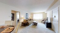 This single-floor space centers around a generously sized living room that offers a warm and inviting atmosphere. The room features wall-to-wall beige carpeting that adds a soft, comfortable feel underfoot, complemented by neutral-toned walls that create a bright and airy environment. A large window fitted with vertical blinds serves as the focal point of the main wall, allowing an abundance of natural light to fill the room while providing privacy control.

The textured ceiling adds subtle visual interest overhead. The living area is well-proportioned, providing ample space for a full seating arrangement, including a cushioned sofa, an accent chair, and both a coffee table and side table in rich wood tones. A television media console is positioned along one wall, offering a practical entertainment setup.

An open doorway leads to an adjacent room, suggesting a connected, flowing floor plan that enhances the sense of space. Wall art and warm-toned lamps contribute to the overall cozy and livable ambiance. The layout and design of this space prioritize comfort and functionality, making it a practical and welcoming living environment.