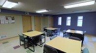 This single-floor space is designed to accommodate group work, training, or classroom-style activities. The room is painted in a bold, deep blue that gives it a distinct and focused atmosphere, contrasted by light-colored flooring with subtle tile accents. Multiple rectangular tables with accompanying chairs are arranged throughout the room, providing flexible seating for a variety of group sizes and configurations. The furniture includes a mix of padded teal-green chairs and standard black-framed chairs, offering comfortable seating options for extended use. Overhead fluorescent lighting ensures the space is well-lit throughout, supplemented by natural light streaming in through windows along one wall. The windows offer a view to the exterior, contributing to an open and airy feel despite the interior setting. Several interior doors along one wall suggest access to additional rooms or utility areas. A small storage unit is also present, adding to the functional nature of the space. Overall, this room is purpose-built for productivity and collaboration, making it well-suited for educational, professional, or community-oriented gatherings.
