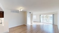 This single-floor space offers a clean, open layout designed to maximize both comfort and functionality. The interior is finished with light-toned hardwood flooring throughout, lending warmth and a sense of continuity across the living areas. Crisp white walls and smooth ceilings enhance the airy, minimalist aesthetic, while recessed HVAC vents integrated into the walls and ceiling ensure a streamlined appearance.

The main living area is generously proportioned, with an open flow that connects seamlessly to an adjoining room or dining area. A partial dividing wall creates a subtle separation between spaces without compromising the open feel. Track lighting on the ceiling provides practical illumination, complementing the abundant natural light that pours in through large floor-to-ceiling windows and sliding glass doors along the far wall. These expansive windows offer views of outdoor greenery, bringing the outside in and creating a bright, welcoming atmosphere.

The overall ambiance is light, calm, and move-in ready, with neutral finishes that provide a versatile backdrop adaptable to a variety of interior styles. The combination of natural light, open space, and quality flooring makes this a well-appointed and inviting single-floor property.