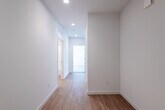 This thoughtfully designed single-floor unit offers a clean, modern aesthetic characterized by crisp white walls, warm light wood-tone flooring, and recessed lighting throughout. The spacious hallway serves as a welcoming corridor, lined with recessed ceiling lights and built-in HVAC vents, leading seamlessly through the unit to a bright entry door.

The open-concept living and kitchen area is a standout feature of the space. A large floor-to-ceiling window floods the room with natural light and provides an outward view, creating an airy and expansive atmosphere. The kitchen is well-equipped with a contemporary two-tone cabinetry design — dark espresso lower cabinets paired with crisp white upper cabinets — complemented by a white quartz countertop, under-cabinet lighting, a stainless steel sink, and a full suite of stainless steel appliances including a refrigerator, dishwasher, range, and microwave.

The bathroom is clean and functional, featuring light wood flooring consistent with the rest of the unit, a vanity with dark cabinetry, a toilet, and a full shower-tub combination. The bedroom offers a softer feel with plush carpeting and a window that brings in natural light, providing a comfortable and restful retreat. The overall ambiance of the space is bright, minimalist, and move-in ready.