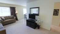 This single-floor space offers a clean and comfortable layout designed for everyday living. The living area is finished with plush wall-to-wall carpeting that adds warmth underfoot, while light-toned walls reflect natural light streaming in through the windows, lending the space an airy, open feel. Dark curtain panels provide a tasteful contrast and allow for flexible light control.

The living area is anchored by a classic dark-wood media console, complete with cabinet storage below, topped by a flat-screen television and a large framed mirror that visually expands the room. A coordinating floor lamp and upholstered armchair round out the seating area, offering a relaxed setting for lounging.

Adjacent to the main living space, a dining nook is visible, furnished with a simple wooden table and cross-back chairs, ideal for casual meals. A baseboard heating system runs along the perimeter of the room, ensuring year-round comfort. Decorative touches, including a potted floral arrangement and framed wall art, add personality without overwhelming the space. Overall, the layout is practical and well-proportioned, making efficient use of its single-floor footprint while maintaining a tidy, inviting ambiance.