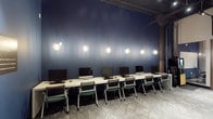 This single-floor computer lab is designed with both functionality and aesthetics in mind. The space is anchored by a long, continuous white workstation surface running along a striking deep-blue accent wall, providing a clean and organized setup for multiple users simultaneously. Each workstation is equipped with a desktop computer and paired with a modern folding chair featuring teal upholstered seats, combining practicality with a touch of color.

Overhead, a series of globe-shaped pendant lights hang at varying heights along a ceiling-mounted track, casting a warm, ambient glow against the dark backdrop of the accent wall. This lighting arrangement serves both a functional and decorative purpose, contributing to the space's contemporary character. The ceiling itself reveals an industrial aesthetic, with exposed structural elements and raw finishes that add depth and texture to the overall design.

The flooring consists of patterned carpet tiles in a neutral gray tone, helping to reduce noise and define the workspace. A substantial structural column is visible along one side of the room, reinforcing the industrial architectural style of the building. Together, these elements create a space that is professional, visually engaging, and well-suited for collaborative or individual computer-based work.