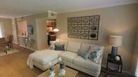 This single-floor residence offers a thoughtfully designed layout that balances comfort and style. The living room serves as the heart of the home, centered around a generous sectional sofa in a soft cream upholstery, accented with decorative pillows in blue and green patterns. A striking geometric wall art piece anchors the main wall, adding a modern yet timeless focal point to the space. Crown molding along the ceiling lends an elegant, finished look to the room, while warm neutral tones on the walls create a cozy and inviting ambiance. The living area flows seamlessly into the kitchen, which is visible through an open passageway. The kitchen features warm wood cabinetry, dark appliances, and pendant lighting that casts a warm glow over the workspace — ideal for both everyday cooking and entertaining. Hardwood-style flooring runs throughout, tying the spaces together cohesively. The overall design aesthetic is casual yet refined, with carefully chosen décor and finishes that reflect a comfortable, livable home. The open sightlines between the living and kitchen areas enhance the sense of space and make the floor plan feel both practical and welcoming.