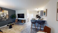 This single-floor unit offers a thoughtfully designed open-concept layout that seamlessly connects the living area and kitchen. The living space features light wood-style flooring throughout, a bold dark accent wall that serves as an eye-catching focal point, and ample room for comfortable furnishings. A wall-mounted flat-screen television anchors the living area, complemented by warm, modern décor elements.

The kitchen is efficiently arranged with gray shaker-style cabinetry, white countertops, and a tiled backsplash. A peninsula with bar seating for two provides a casual dining option and reinforces the open flow between the kitchen and living space. Stainless steel appliances, including a refrigerator and microwave, complete the kitchen's functional design.

The bedroom is a calm, neutral-toned retreat featuring the same light wood-style flooring. A wide horizontal window frames lush greenery and palm foliage outside, allowing natural light to fill the room. An open closet provides organized storage space, and an adjoining bathroom is conveniently accessible directly from the bedroom. The overall ambiance of the unit is clean, contemporary, and inviting.