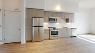 This single-floor unit showcases a clean, contemporary design with consistent light wood flooring throughout the main living areas. The kitchen is a standout feature, outfitted with flat-front cabinetry in a warm taupe finish, contrasted by white subway tile backsplash and dark countertops. Stainless steel appliances include a full-size refrigerator, range with oven, built-in microwave, and dishwasher, providing a fully functional cooking space. Ample upper and lower cabinetry offers generous storage.

The bathroom continues the modern aesthetic with white cabinetry, a dual-sink vanity topped with light stone counters, and a large mirror with elegant globe lighting above. A soaking tub with integrated shower fixtures sits adjacent, while light wood flooring adds warmth. A walk-in closet is directly accessible from the bathroom, providing convenient storage.

The bedroom is bright and airy, benefiting from a large window positioned to capture natural light and offer open outdoor views. Neutral carpet, crisp white walls, and a ceiling fan complete the comfortable, well-lit space. The overall layout, visible in the floor plan, is efficiently organized with a flowing open design.