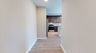 This well-appointed single-floor unit offers a thoughtfully designed open-concept layout with consistent light wood-tone flooring throughout, creating a seamless and cohesive living environment. The kitchen serves as a central highlight, showcasing rich dark walnut cabinetry, granite countertops with subtle speckled detailing, and a full suite of stainless steel appliances including a refrigerator, range, microwave, and dishwasher. Track lighting and elegant pendant fixtures illuminate the space, while a central island with matching granite provides additional prep and gathering space. The living area flows naturally from the kitchen, benefiting from abundant natural light streaming through large windows that frame open outdoor views of the surrounding landscape and skyline. The bedrooms are generously proportioned, each featuring oversized windows that flood the rooms with daylight and offer elevated views of the neighborhood and greenery beyond. The bathroom is finished with the same dark cabinetry found in the kitchen, complemented by a granite-topped vanity, a large mirror, and ample storage. Neutral warm-toned walls throughout the unit contribute to a calm, modern ambiance, making this a well-balanced and functional living space.