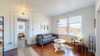 This single-floor residence artfully combines historic architectural character with contemporary design sensibilities. Rich hardwood floors flow through the main living areas, anchoring a bright and airy living room fitted with large street-facing windows that invite generous natural light and neighborhood views. A cast-iron radiator adds a period-appropriate accent alongside a textured ceiling and classic door moldings that speak to the building's heritage.

The bedroom spaces offer distinct personalities: one features dramatic exposed brick walls on all sides, creating a cozy, loft-like atmosphere with an undeniable urban charm, while a second bedroom is bright and minimal, with a window overlooking the streetscape and a classic radiator for warmth.

The bathroom is a standout, centered around a striking clawfoot soaking tub with a ceiling-mounted shower, tiled flooring in a neutral slate tone, and a window that keeps the space light and fresh. A vintage-style radiator completes the room's classic character.

The kitchen is practical and well-appointed, featuring rich wood cabinetry, granite-look countertops, a stainless undermount sink, and a full-size refrigerator. A hallway with an accessible electrical panel connects the rooms efficiently. Throughout the space, the interplay of original architectural details and thoughtful modern furnishings creates a welcoming and functional home.
