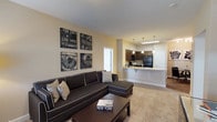 This single-floor residence offers a thoughtfully designed open-concept layout that seamlessly connects the living, dining, and kitchen areas. The living room is anchored by plush carpeting, a large sectional sofa, and a private exterior door that allows natural light to filter in through a window with blinds. The kitchen is well-equipped with rich dark wood cabinetry, granite countertops, black appliances including a gas range, microwave, and refrigerator, and a breakfast bar peninsula illuminated by stylish pendant lighting. Wood-look flooring runs through the kitchen and dining area, where a compact dining set sits adjacent to decorative wall art. Both bathrooms feature dark wood vanities with granite countertops, wood-look flooring, and a tub-shower combination. One bathroom also includes built-in wall shelving for added storage. The bedroom is carpeted and filled with natural light from a window dressed in patterned curtains. A generously sized walk-in closet with wire shelving and hanging rods provides ample storage. Throughout the residence, neutral warm-toned walls and consistent finishes create a cohesive and welcoming ambiance.