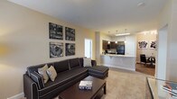 This single-floor residence offers a thoughtfully designed open-concept layout that seamlessly connects the living, dining, and kitchen areas. The living room is anchored by plush carpeting, a large sectional sofa, and a private exterior door that allows natural light to filter in through a window with blinds. The kitchen is well-equipped with rich dark wood cabinetry, granite countertops, black appliances including a gas range, microwave, and refrigerator, and a breakfast bar peninsula illuminated by stylish pendant lighting. Wood-look flooring runs through the kitchen and dining area, where a compact dining set sits adjacent to decorative wall art. Both bathrooms feature dark wood vanities with granite countertops, wood-look flooring, and a tub-shower combination. One bathroom also includes built-in wall shelving for added storage. The bedroom is carpeted and filled with natural light from a window dressed in patterned curtains. A generously sized walk-in closet with wire shelving and hanging rods provides ample storage. Throughout the residence, neutral warm-toned walls and consistent finishes create a cohesive and welcoming ambiance.