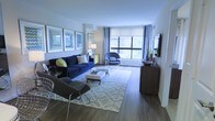 This single-floor space centers around a thoughtfully designed living area that combines contemporary style with functional comfort. The room features warm hardwood flooring that extends throughout, providing a cohesive and polished foundation. Large windows along one wall allow generous amounts of natural light to fill the space, offering views of lush greenery outside and creating an open, airy atmosphere.

The living area is anchored by a bold navy blue sofa accented with decorative pillows, complemented by a coordinating accent chair and a modern lounge chair, together forming an inviting conversational layout. A geometric-patterned area rug in neutral tones defines the seating zone and adds visual texture underfoot. A sleek, low-profile white coffee table serves as a practical centerpiece.

Wall-mounted framed artwork adds a curated, gallery-like quality to the space, while a floor lamp and a decorated console table provide both ambient lighting and stylish storage. A wooden media console along one wall offers additional storage and display space. The overall ambiance is modern and inviting, balancing cool tones with warm wood finishes throughout.