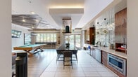 Located at 301 Continental Parkway in Newport News, Virginia, this single-floor community lounge space offers a well-appointed and visually dynamic environment designed for socializing and relaxation. The open-concept layout connects a fully equipped kitchen with a comfortable living area, creating a seamless flow throughout the space.

The kitchen is a standout feature, showcasing warm walnut cabinetry that extends floor to ceiling, complemented by a striking gold mosaic tile backsplash that serves as a bold focal point. A clean white countertop runs the length of the wall, housing a stainless steel sink, ice maker, undercounter refrigerator, and built-in microwave. Stylish globe pendant lights hang above, adding a modern, polished touch. Large-format light tile flooring grounds the kitchen area and extends into the hallway.

The living area transitions to rich hardwood flooring and is anchored by a vibrant teal accent wall featuring two wall-mounted flat-screen televisions above a decorative media console. Brown leather armchairs, organic live-edge coffee tables, and a plush area rug contribute to a warm yet contemporary atmosphere. A billiards table adds a recreational focal point to the room. Curated triptych landscape artwork adorns the walls, and a sculptural multi-globe chandelier provides elegant overhead lighting. Natural light filters in through large windows, enhancing the space's inviting ambiance.