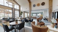 This thoughtfully designed clubhouse offers a welcoming, open-concept layout that seamlessly blends comfort and modern style. The space is anchored by dramatically soaring ceilings supported by exposed structural columns, creating an airy, grand atmosphere throughout the main gathering area. Floor-to-ceiling windows flood the interior with natural light while framing views of the surrounding community and outdoor amenities.

The lounge area features a mix of plush seating, including a cream sofa, tufted ottoman, and wingback accent chairs in rich cognac leather, all arranged to encourage relaxed conversation. Decorative wall art and lush indoor plants add warmth and texture to the neutral palette. Adjacent dining and high-top seating areas are furnished with contemporary bistro and counter-height tables, ideal for casual gatherings or individual use.

The fully equipped kitchen is a standout feature, showcasing warm wood cabinetry, a large white quartz island with bar seating, a textured mosaic tile backsplash, and a series of striking pendant lights overhead. A dedicated coffee and beverage station, microwave, and dishwasher make the space highly functional for community events. Large windows throughout ensure the kitchen remains bright and connected to the outdoors. The large-format tile flooring ties the entire space together with a clean, cohesive finish.