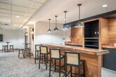 Located at 3841 E Washington Ave in Madison, Wisconsin, this single-floor amenity space offers a generous footprint designed to accommodate both active and social pursuits. The layout is anchored by a broad, carpeted hallway that connects each area seamlessly, featuring recessed lighting, drop ceilings, warm wood baseboards, and tasteful wall art that creates a welcoming first impression.

The living room lounge is a standout gathering space, highlighted by a stylish bar counter with dark cabinetry, warm wood accents, and pendant lighting overhead. Bar-height seating lines the counter, while open floor space accommodates multiple seating arrangements, including round tables and chairs. A wall-mounted television adds an entertainment element, making this area well-suited for social gatherings.

The exercise room is generously proportioned and thoughtfully outfitted with cardio equipment including treadmills and elliptical machines, a multi-station cable weight machine, a dumbbell rack, and stability balls. Natural light filters in through multiple windows, complementing the overhead panel lighting and durable rubberized flooring. A two-tone wall treatment in blue and cream adds a clean, energetic feel to the space.

Throughout, the design maintains a cohesive, professional aesthetic with neutral tones, carpeted corridors, and glass-paneled interior doors that enhance both flow and visibility across the floor.