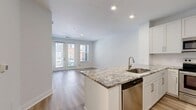 This 690-square-foot, one-bedroom, one-bathroom apartment at 580 S Goddard Blvd is designed with modern finishes and an efficient, open layout. Light wood-tone flooring flows consistently throughout the entire unit, creating a cohesive and airy feel. The combined kitchen and living area serves as the heart of the home, anchored by a striking granite-topped kitchen island with an undermount sink. The kitchen features white shaker cabinetry, granite countertops, and a full suite of stainless steel appliances including a range, microwave, and refrigerator. A dedicated in-unit laundry closet with a stacked washer and dryer adds everyday convenience. The living area is bathed in natural light through glass doors that open to an outdoor space, complemented by an additional side window with views of the surrounding landscape. The 170-square-foot bedroom includes a ceiling fan, a large window overlooking greenery, and direct access to a closet and hallway. The bathroom offers a tiled tub-shower combination with white subway tile, marble-look tile flooring, brushed gold fixtures, and a granite vanity countertop. A dedicated hallway connects all rooms, completing this well-appointed and functional apartment.