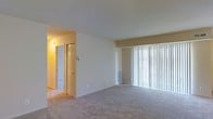 This single-floor residence at 7916/302 offers a clean, well-maintained layout designed for comfortable everyday living. The main living area is a standout feature, characterized by soft wall-to-wall carpeting in neutral tones that adds warmth underfoot. A large floor-to-ceiling vertical blind-covered sliding glass door floods the space with natural light and provides an open, airy feel throughout the room. The walls are finished in a light beige tone, contributing to a calm and neutral ambiance that suits a variety of interior styles. A wall-mounted heating vent is positioned beneath the large window, ensuring efficient climate comfort in the living space. Adjacent to the main room, a hallway leads to additional areas of the unit, with a classic paneled closet door visible, hinting at ample storage options. The hallway is warmly lit, offering a welcoming transition between spaces. The overall design is straightforward and functional, with clean lines, smooth ceilings, and a practical flow that makes the space easy to navigate. The unit presents a move-in ready condition with its neutral finishes and well-preserved surfaces throughout.