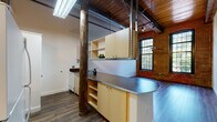 Located at 51 Front St in Cumberland, Rhode Island, this loft-style unit occupies a thoughtfully converted historic mill building, where original industrial character meets contemporary design. Throughout the space, rich hardwood-style flooring flows seamlessly underfoot, complementing the warmth of the wood-plank ceilings overhead. Exposed original timber posts add authenticity and structural charm, while visible ductwork and track lighting reinforce the industrial aesthetic.

The open-plan living area is anchored by a striking wall of exposed brick, punctuated by large arched windows with multi-pane glazing that draw in generous natural light and offer views of surrounding greenery. A ceiling fan adds both function and character to this expansive, airy room.

The kitchen is integrated into the open layout via a peninsula featuring sleek stainless steel countertops and light wood cabinetry, providing both workspace and a natural divide between cooking and living areas. Appliances include a white range, over-the-range microwave, and a tile backsplash, with upper open shelving for display and storage.

The bathroom is well-appointed with a wide vanity, warm-toned countertop, large mirror, and multi-bulb light fixture, creating a bright and functional space. The overall ambiance throughout is one of industrial-chic character, combining the heritage of its mill origins with clean, modern livability.