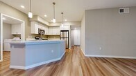 This thoughtfully designed 3-bedroom, 2-bathroom single-story residence offers a functional and modern living experience. The open-concept kitchen and living area serves as the heart of the home, featuring warm wood-look flooring, a curved light blue kitchen island, crisp white cabinetry, stainless steel appliances including a refrigerator and microwave, and stylish pendant lighting overhead. Recessed lighting further brightens the space throughout.

The primary bedroom is generously sized, complete with a ceiling fan, sliding glass doors that open to a private outdoor balcony, and abundant natural light. It is complemented by a walk-in closet outfitted with dual clothing rods and shelving on carpet flooring. The secondary bedrooms also feature plush carpet and ample natural light from well-placed windows.

Both bathrooms are finished with white cabinetry, stone-topped vanities, large mirrors, and wood-look tile flooring. One bathroom includes a full tub-shower combination. A dedicated laundry room houses a full-size washer and dryer with overhead storage cabinets and direct exterior door access. Hallways connect the spaces efficiently, and neutral warm-gray wall tones create a cohesive, calm ambiance throughout the entire residence.