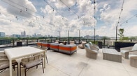 Perched atop 400 Bishop St NW in Atlanta's dynamic West Midtown neighborhood, this rooftop terrace offers an impressive open-air retreat with sweeping panoramic views of the Atlanta skyline and surrounding greenery. The generously sized outdoor deck features large-format light-toned concrete pavers that create a clean, modern foundation throughout the space.

The terrace is thoughtfully arranged with multiple seating areas, including plush gray and orange modular sofas, woven rattan lounge chairs with soft cushions, and bistro-style dining tables with woven chairs — providing versatile options for both relaxed lounging and social gatherings. Tall planters with lush greenery add natural accents and subtle privacy between zones.

Overhead, strands of cafe-style string lights crisscross the open sky, lending a warm, inviting ambiance ideal for evening use. A sleek metal railing system lines the perimeter, ensuring unobstructed sightlines across the cityscape.

A modern interior lounge area is accessible through large floor-to-ceiling glass sliding doors and double glass-pane entry doors framed in dark bronze, creating a seamless visual and physical connection between the indoor and outdoor spaces. The interior portion features track lighting and additional dining furnishings, extending the functionality of the space year-round. Together, the indoor and outdoor areas form a cohesive, well-appointed rooftop amenity ideal for socializing and relaxation.