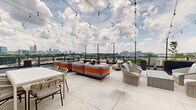 Perched atop 400 Bishop St NW in Atlanta's dynamic West Midtown neighborhood, this rooftop terrace offers an impressive open-air retreat with sweeping panoramic views of the Atlanta skyline and surrounding greenery. The generously sized outdoor deck features large-format light-toned concrete pavers that create a clean, modern foundation throughout the space.

The terrace is thoughtfully arranged with multiple seating areas, including plush gray and orange modular sofas, woven rattan lounge chairs with soft cushions, and bistro-style dining tables with woven chairs — providing versatile options for both relaxed lounging and social gatherings. Tall planters with lush greenery add natural accents and subtle privacy between zones.

Overhead, strands of cafe-style string lights crisscross the open sky, lending a warm, inviting ambiance ideal for evening use. A sleek metal railing system lines the perimeter, ensuring unobstructed sightlines across the cityscape.

A modern interior lounge area is accessible through large floor-to-ceiling glass sliding doors and double glass-pane entry doors framed in dark bronze, creating a seamless visual and physical connection between the indoor and outdoor spaces. The interior portion features track lighting and additional dining furnishings, extending the functionality of the space year-round. Together, the indoor and outdoor areas form a cohesive, well-appointed rooftop amenity ideal for socializing and relaxation.