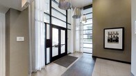 This single-floor space presents a polished and contemporary lobby entrance designed to make a strong first impression. The entryway is defined by dramatic floor-to-ceiling glass curtain walls framed in sleek black metal, flanking a set of glass double doors that flood the interior with abundant natural light. Sheer white drapery panels line the expansive windows, softening the light and adding an elegant, refined touch to the space.

Overhead, a series of large metallic pendant lights hang at varying heights, serving as a bold architectural statement while providing warm, focused illumination throughout the entry corridor. The ceiling height is notably generous, contributing to an open and airy sense of scale.

The flooring transitions from large-format neutral tiles to a textured entry mat at the threshold, combining practicality with a clean aesthetic. An olive-toned accent wall adds depth and warmth to the palette, complementing the cool metallic and glass elements throughout.

Overall, the space strikes a careful balance between modern commercial design and an inviting ambiance. The interplay of natural light, high ceilings, transparent facades, and carefully chosen materials creates a lobby that is both functional and visually impressive, setting a professional and welcoming tone for all who enter.