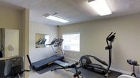 This single-level fitness room provides a practical and well-equipped space for cardiovascular exercise. The room features neutral beige walls, a textured ceiling with fluorescent lighting that ensures bright, even illumination throughout. A window along one wall allows natural light to filter in, offering a view of the surrounding greenery outside. The space is outfitted with multiple treadmills, an elliptical machine, and a weight bench, providing a variety of workout options. A wall-mounted mirror on one side visually expands the room while allowing users to monitor their form during exercise. The flooring is covered with dark rubber mats beneath the equipment, offering both protection and stability. A security camera is mounted near the ceiling, adding a layer of safety to the space. The overall layout is efficient, making use of the available area to accommodate several machines without feeling overly cramped. The clean, minimalist design keeps the focus on functionality, making this fitness room a straightforward and accessible option for those looking to maintain an active lifestyle.