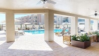 This expansive outdoor amenity space is centered around a sparkling, resort-style swimming pool framed by a generously sized pool deck finished in light-toned paver stone. Rows of lounge chairs and patio furniture are arranged throughout the sundeck, offering ample space for relaxation and recreation under the open sky. A large covered patio area extends along one side of the pool, supported by sturdy stucco columns and equipped with multiple ceiling fans to provide comfortable shade and airflow. Beneath this pergola-style structure, thoughtfully arranged seating groups — composed of wood-framed chairs and sofas with clean, neutral upholstery — are nestled alongside oversized rectangular planters filled with lush greenery, adding a natural, resort-like ambiance. Market umbrellas are positioned throughout the open deck for additional sun coverage. The surrounding architecture features a warm, Southwestern-influenced stucco exterior with multi-story residential buildings rising in the background. Large glass doors connect the covered lounge area to an adjacent interior amenity space, enhancing the indoor-outdoor flow. The overall atmosphere is bright, airy, and well-appointed, offering a polished communal retreat designed for both leisure and socializing.