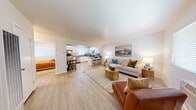 This single-floor residence offers a well-designed layout that balances open communal living with comfortable private spaces. The heart of the home is a spacious open-concept living and kitchen area unified by light wood-tone flooring and crisp white walls that create a clean, airy atmosphere. The living room accommodates generous seating and is brightened by a large window with blinds, while a bold coastal-inspired art piece anchors the main wall.

The kitchen is thoughtfully appointed with white shaker-style cabinetry, gray countertops, and stainless steel appliances including a gas range with over-the-range microwave. A peninsula with bar seating seamlessly connects the kitchen to the living area, making it ideal for both meal preparation and casual entertaining. Adjacent to the kitchen, a dedicated dining nook provides an intimate space for sit-down meals.

The primary bedroom is a serene retreat, featuring wood-tone flooring, a mirrored closet, and a window that allows natural light to filter in. A second bedroom offers a cozy, versatile space suitable as a guest room or home office. The full bathroom is finished with wood-look flooring, a gray vanity countertop, and a tub-shower combination with a window for natural ventilation. Throughout, the warm, neutral palette and natural wood accents lend the space a cohesive, welcoming character.